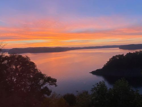 A serene sunset over a calm lake, with vibrant orange and purple hues reflecting on the water and silhouetted trees.