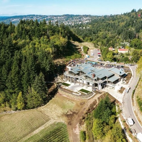 Aerial view of a large building surrounded by trees and hills, with a road and parking area nearby.