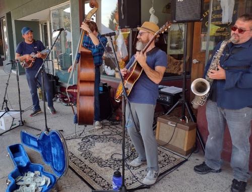 A street band performs with a guitar, saxophone, mandolin, and double bass, surrounded by a crowd and a money case.