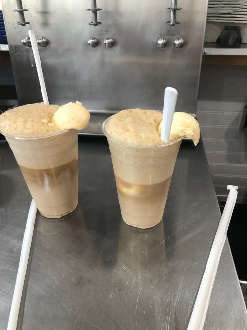 Two frothy ice cream floats in clear cups, with straws, on a stainless steel counter.