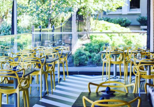 Bright restaurant interior with yellow chairs, glass walls, and striped flooring, surrounded by greenery.