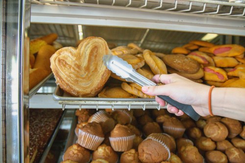 A hand using tongs to pick up a heart-shaped pastry from a tray of assorted baked goods.