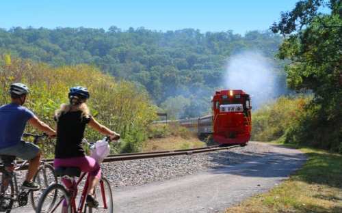 A red train approaches on tracks beside a bike path, with two cyclists enjoying a scenic ride in a lush green landscape.