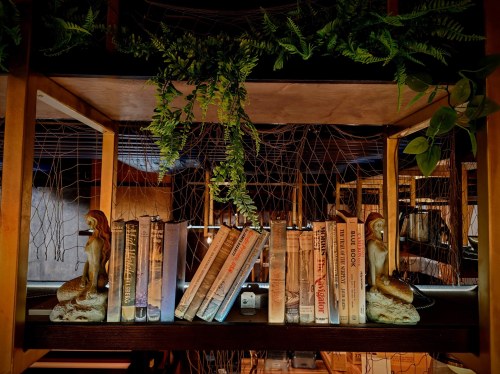 A cozy bookshelf with vintage books, adorned with greenery and mermaid sculptures on either side.