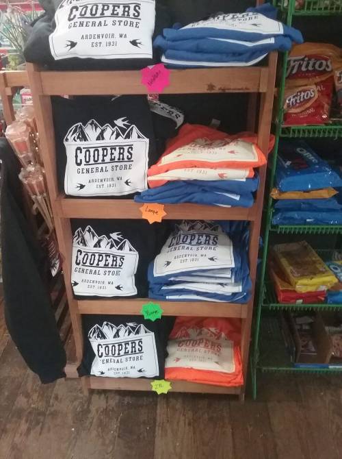 A wooden shelf displaying folded t-shirts with "Coopers General Store" logo in various colors and sizes.