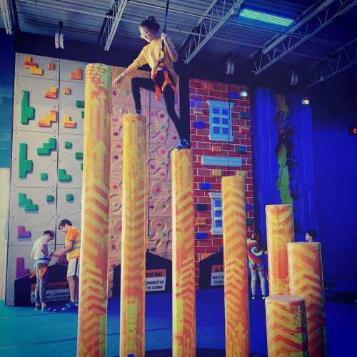 A person climbs colorful vertical pillars in a climbing gym, with others training on a nearby wall.