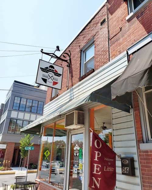 A brick storefront with a sign reading "Welcome to Lititz" and an open flag, featuring nearby modern buildings.