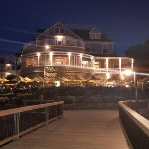 A waterfront restaurant illuminated at night, featuring a large deck and outdoor seating, surrounded by greenery.