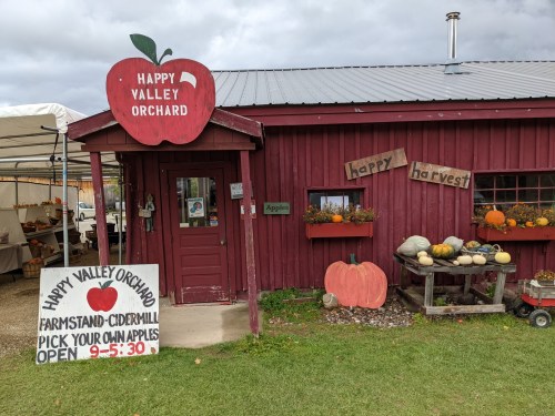 A red barn-style building with a sign for Happy Valley Orchard, featuring pumpkins and a farmstand outside.