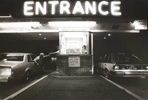 A vintage night scene of a ticket booth at an entrance, with cars parked outside and a glowing sign above.