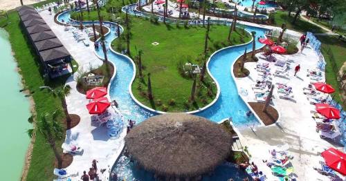 Aerial view of a resort pool area with winding lazy river, palm trees, and lounge chairs under red umbrellas.