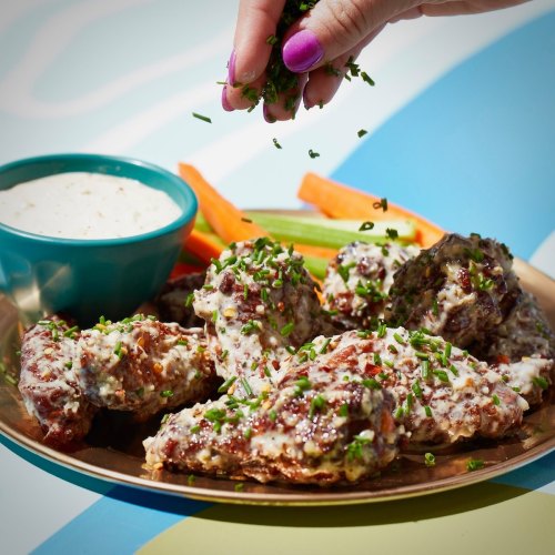 A hand sprinkles herbs over a platter of chicken wings, served with a dipping sauce and fresh vegetables.