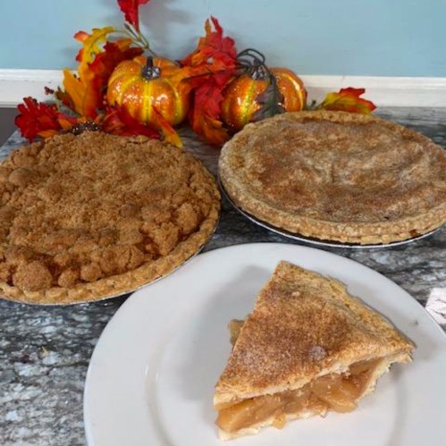 Two pies, one with a crumb topping and one with a golden crust, alongside a slice of apple pie on a plate, with autumn decor.