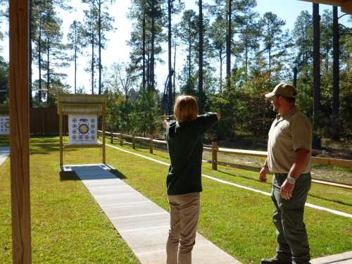 A person aims an arrow at a target while an instructor observes in a wooded outdoor archery range.