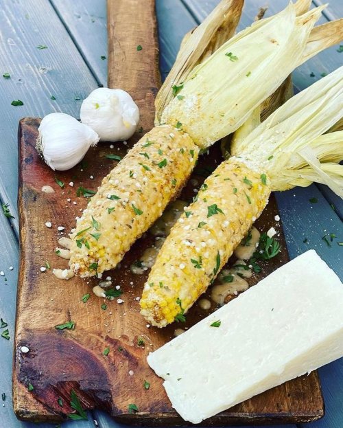 Two ears of corn topped with cheese and herbs, accompanied by garlic and a wedge of cheese on a wooden cutting board.