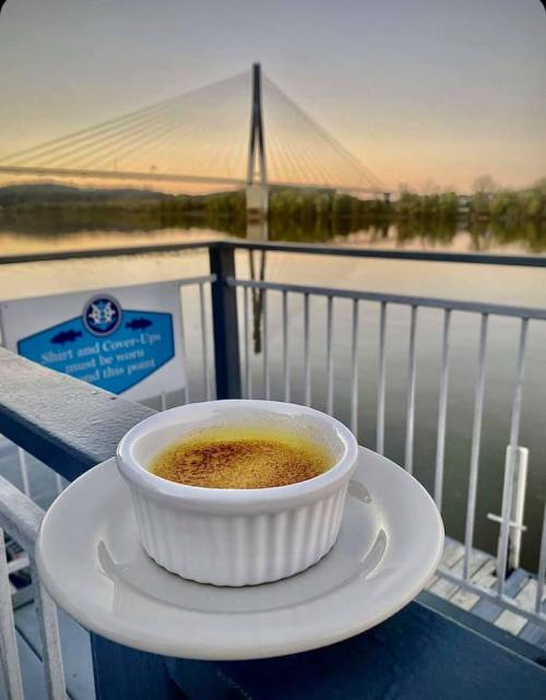 A ramekin of crème brûlée sits on a table with a river and a bridge in the background during sunset.