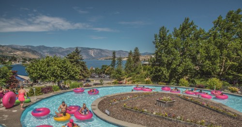 A scenic pool area with colorful inner tubes, surrounded by greenery and mountains in the background under a clear sky.