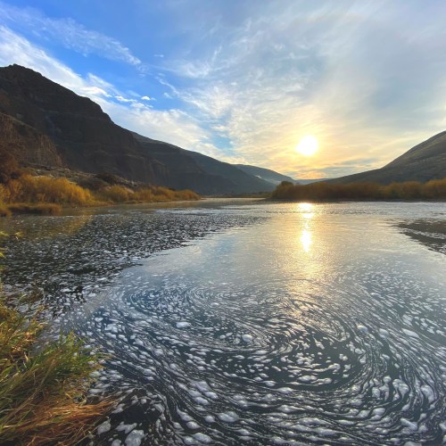 A serene landscape featuring a river reflecting the sunset, surrounded by mountains and lush vegetation.