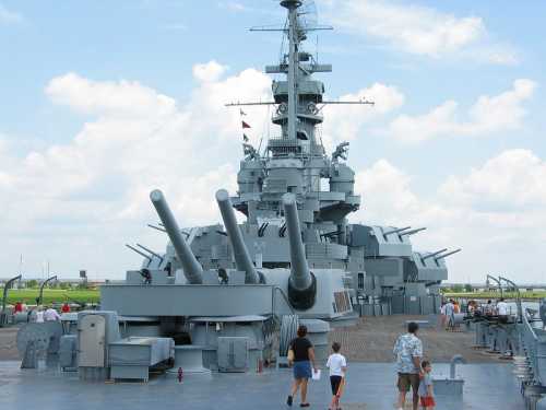 A large battleship with prominent cannons, viewed from the deck, with people walking nearby under a cloudy sky.