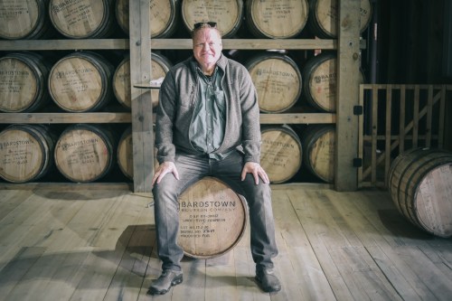 A man sits on a barrel in a distillery, surrounded by stacked barrels in a rustic wooden setting.