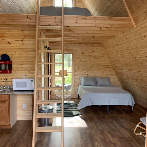 Cozy wooden cabin interior with a lofted bed, kitchenette, and large windows letting in natural light.