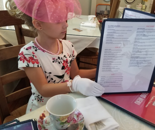 A young girl in a floral dress and pink hat examines a menu at a tea party, wearing gloves and a pearl necklace.