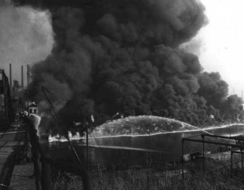 A large plume of black smoke rises from a fire near a body of water, with flames visible in the foreground.