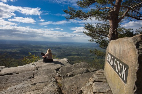 A person sits on a rocky ledge overlooking a vast landscape, with a sign reading "Bald Rock" nearby.