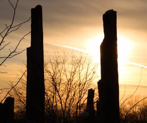 Silhouettes of tall stone pillars against a sunset sky with wispy clouds and bare trees in the foreground.