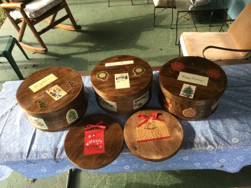 Five wooden boxes with festive decorations and labels, arranged on a blue tablecloth outdoors.