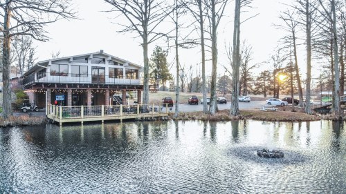 A lakeside building with a deck, surrounded by trees, reflecting in the water at sunset.