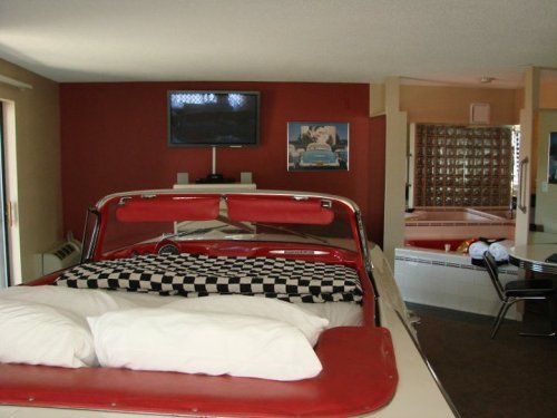 A retro-themed room featuring a bed in a vintage car, a TV on the wall, and a jacuzzi in the background.