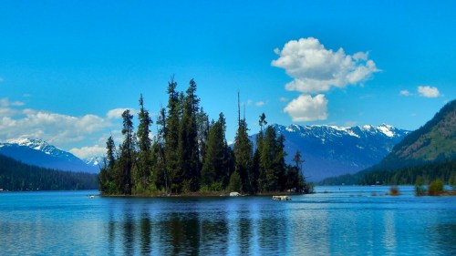 A serene lake scene featuring a small island with trees, surrounded by mountains and a clear blue sky with clouds.