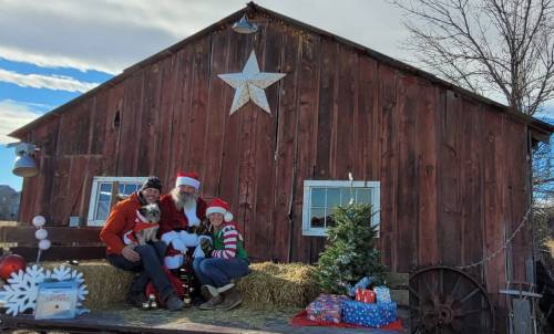A festive scene with Santa, two children, and a dog in front of a rustic barn, decorated for Christmas.