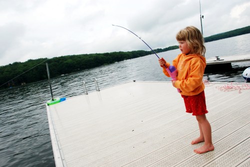 A young girl in an orange hoodie stands on a dock, fishing with a rod over a calm lake.