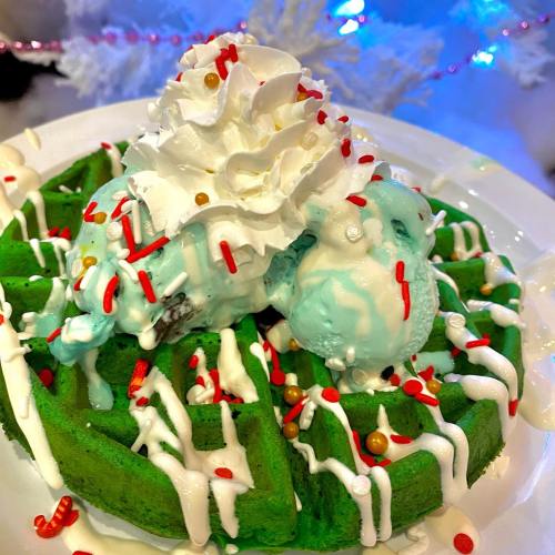 A festive green waffle topped with whipped cream, mint ice cream, and colorful sprinkles, set against a holiday backdrop.