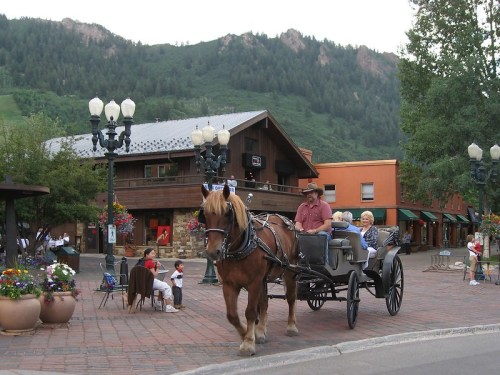 A horse-drawn carriage with a driver and passenger moves through a quaint town square surrounded by shops and mountains.