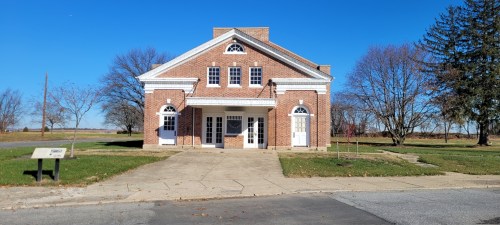 A brick building with white trim and multiple doors, surrounded by grass and trees under a clear blue sky.