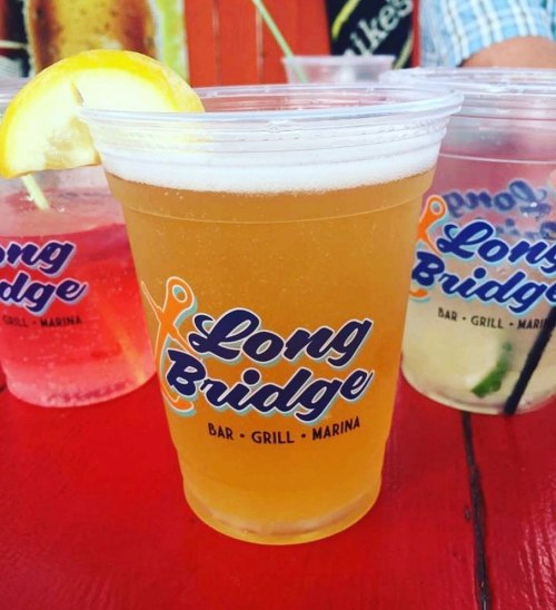 A close-up of a drink in a clear cup with a lemon slice, featuring the "Long Bridge Bar Grill Marina" logo.