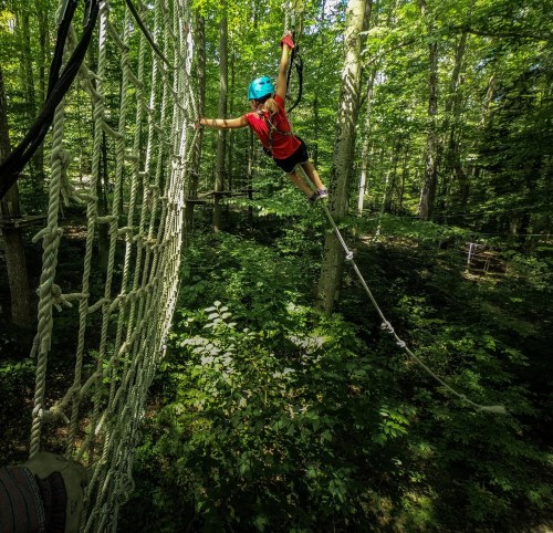 A person in a helmet balances on a rope while reaching for a net in a lush green forest.