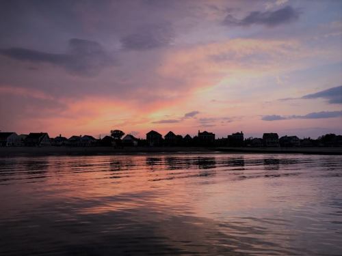 A serene sunset over a calm water body, reflecting colorful clouds and silhouettes of houses along the shore.