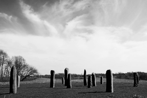 A circle of tall, standing stones in a grassy field under a cloudy sky, with sparse trees in the background.