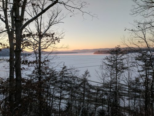 A serene winter landscape featuring a frozen lake at sunset, framed by bare trees and distant mountains.