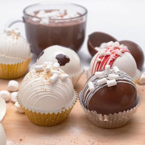 Assorted chocolate bombs topped with marshmallows, alongside a cup of hot chocolate and mini marshmallows.