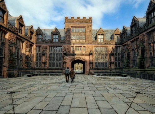Two people walk towards a grand, historic building with intricate architecture under a partly cloudy sky.