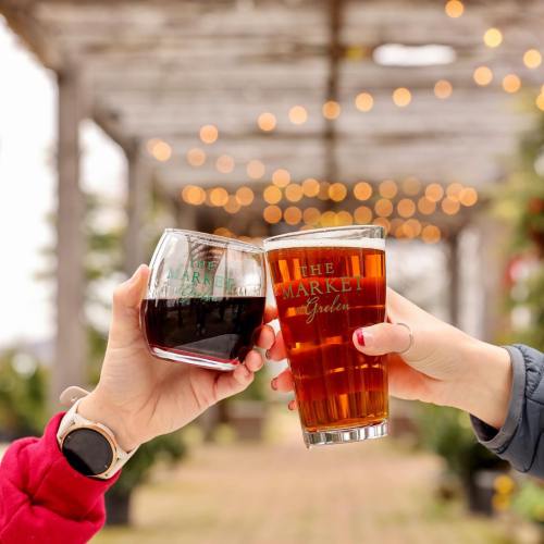 Two people clink glasses of dark and light beverages outdoors, with festive lights in the background.