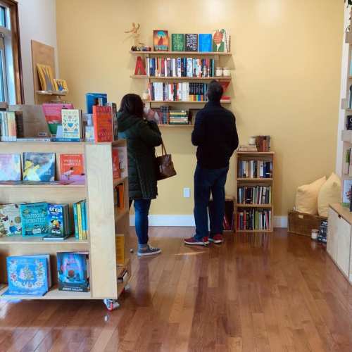 Two people browse bookshelves in a cozy bookstore with wooden floors and colorful displays.