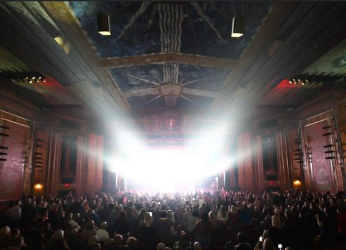 A crowded theater with bright stage lights illuminating the audience and ornate ceiling details.