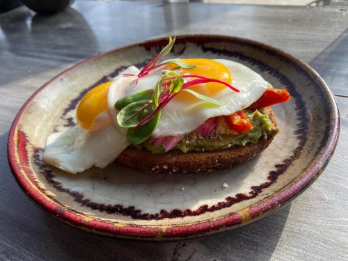 A plate with avocado toast topped with poached eggs, red peppers, and microgreens on a rustic dish.