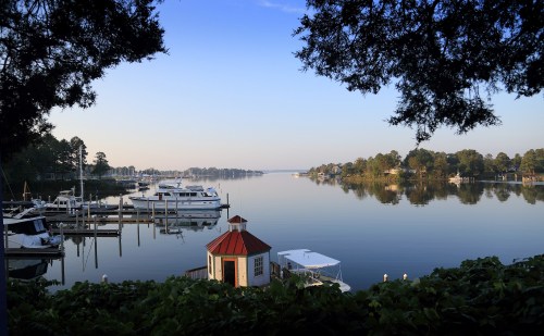 A serene lakeside view with boats docked, surrounded by trees and a small red-roofed building in the foreground.